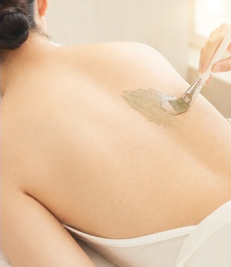 베이지 톤 시술실에서 엎드린 여성의 등에 시술자가 넓은 브러시로 초록빛 해초 필링 제형을 도포하고 있다