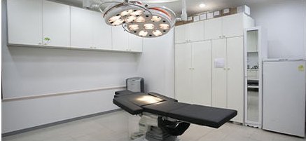 無影灯の付いた施術室の内部 — 黒い施術ベッドと白い収納棚、小型冷蔵庫と鏡台が置かれた様子